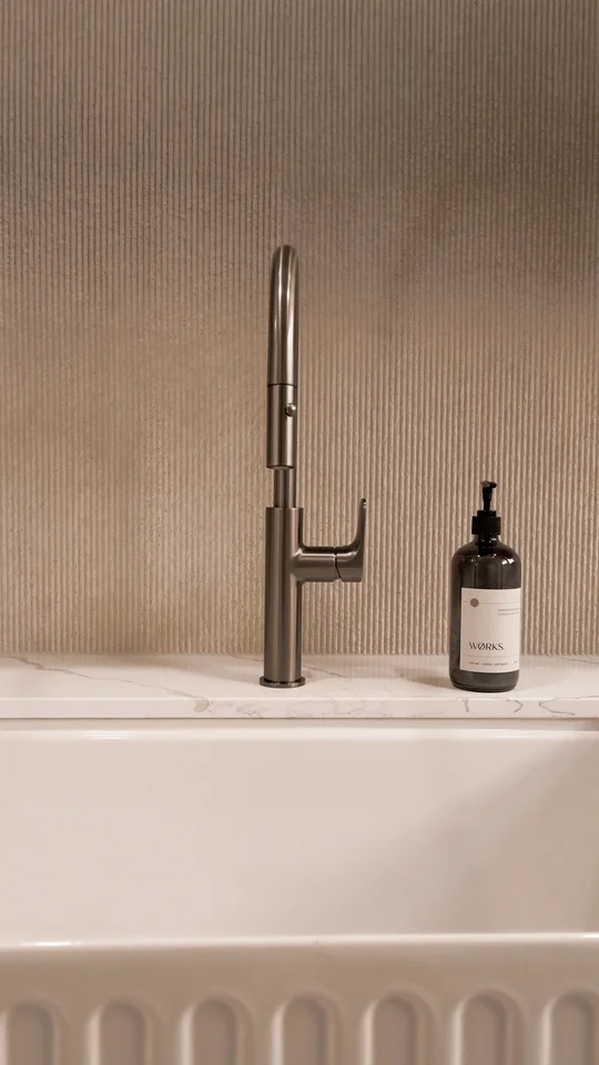 Minimal sink and tall tap with a soap bottle against a textured wall.
