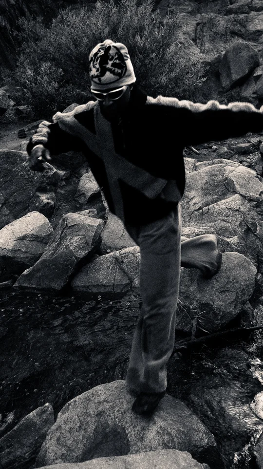 Models wearing black and grey outerwear among rocks and trees.
