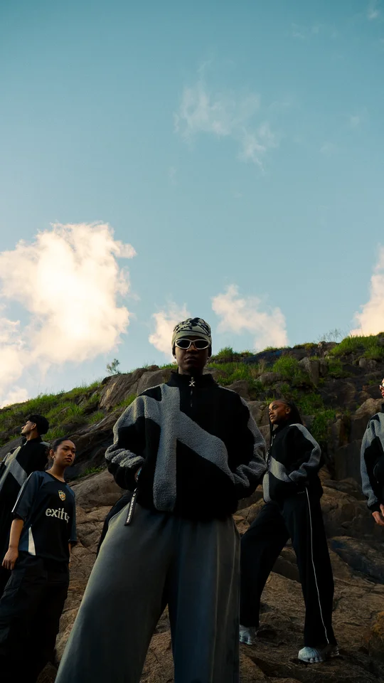 Models in black and grey clothing on a rocky hillside under open sky.