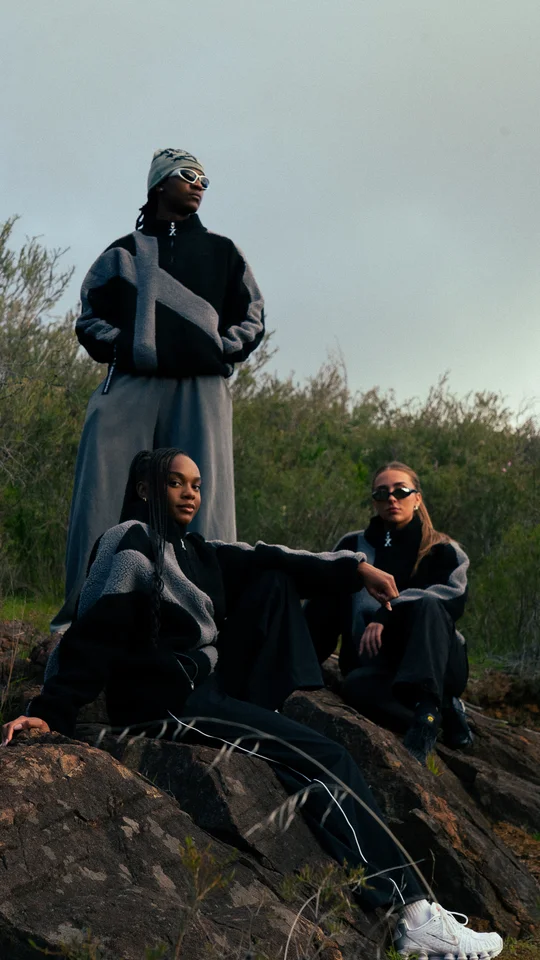 Models in black and grey clothing on a rocky hillside under open sky.