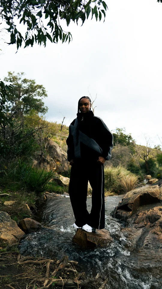 Model in a black and grey jacket standing on rocks beneath a tree.