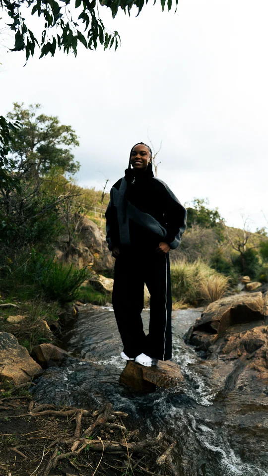 Model in a black and grey jacket standing on rocks beneath a tree.