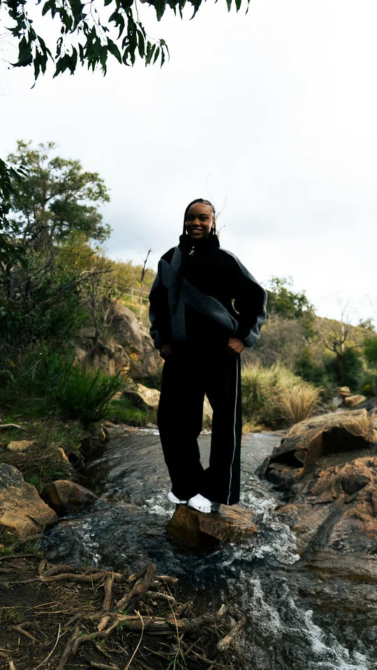 Model in a black and grey jacket standing on rocks beneath a tree.