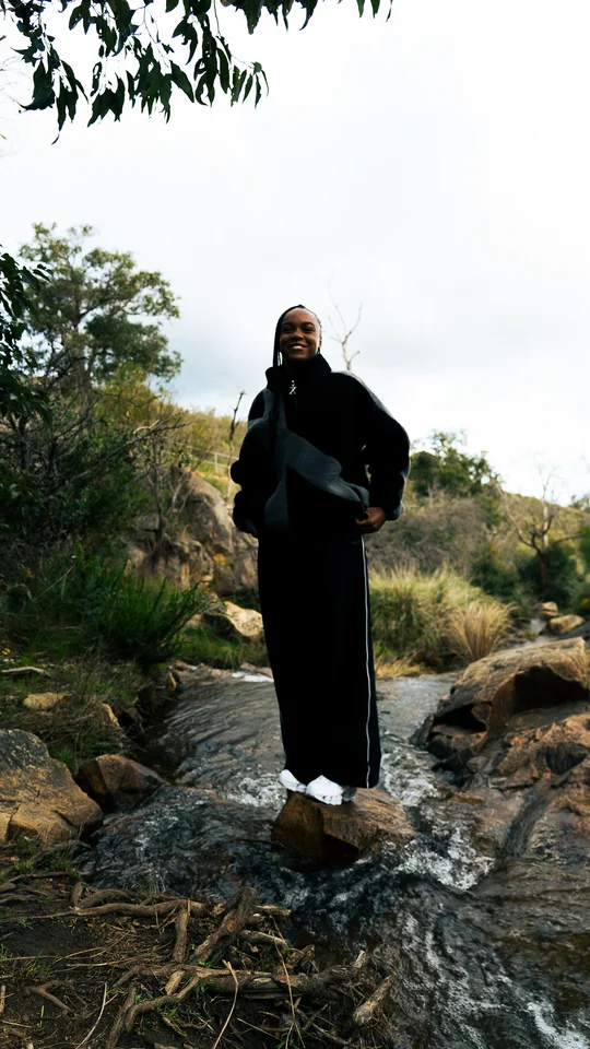 Model in a black and grey jacket standing on rocks beneath a tree.