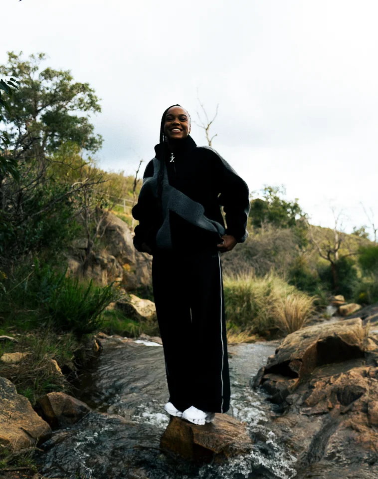 Model in a black and grey jacket standing on rocks beneath a tree.