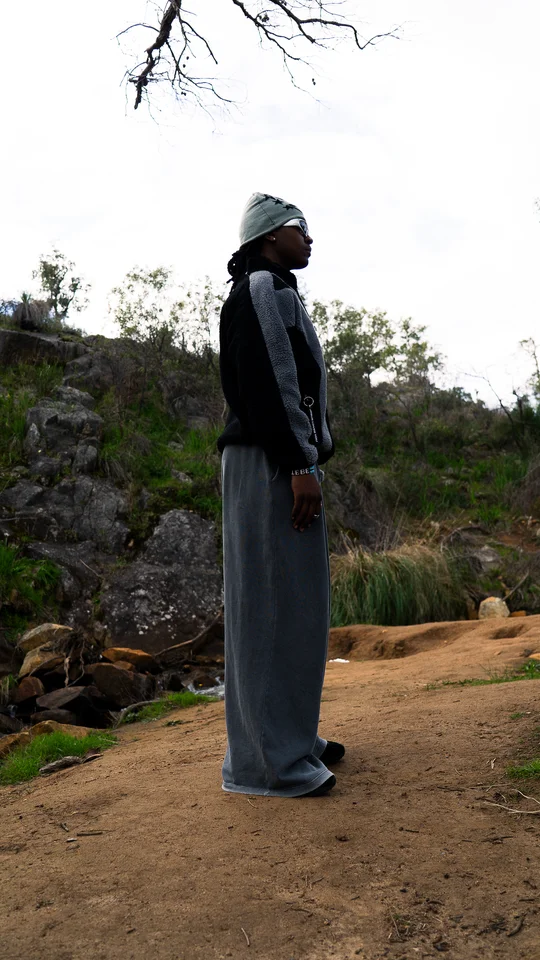 Model wearing a grey beanie, black jacket and wide grey trousers outdoors.
