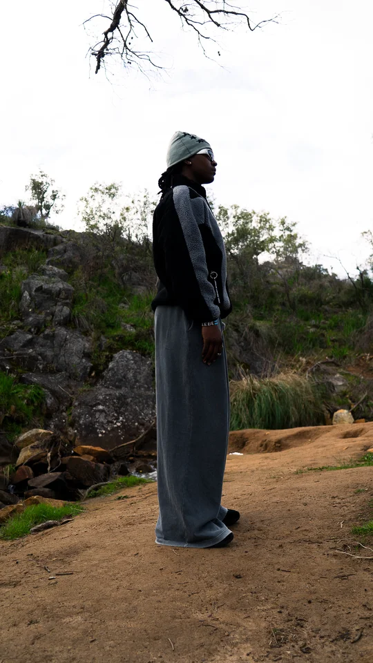 Model wearing a grey beanie, black jacket and wide grey trousers outdoors.