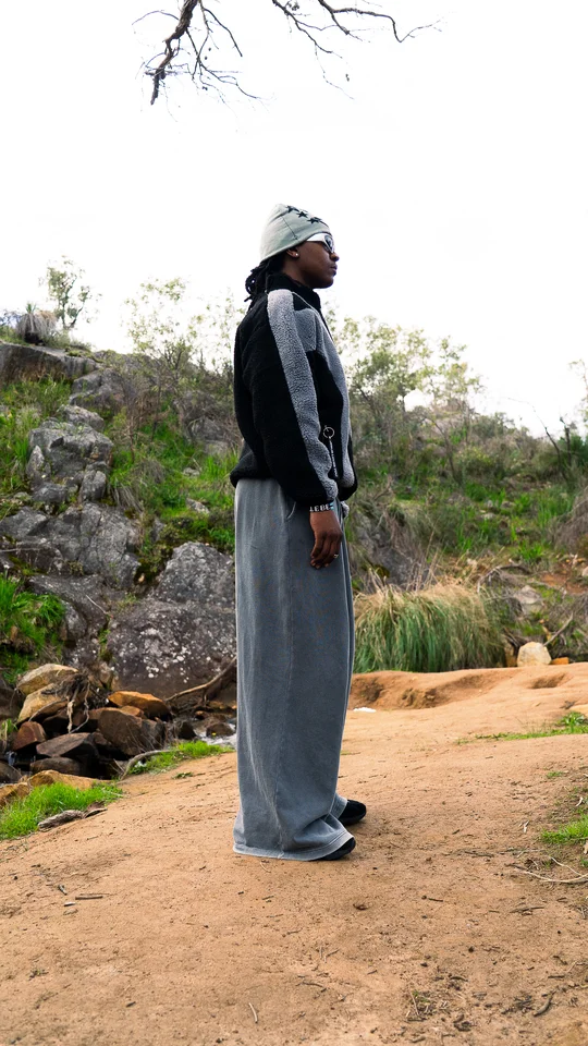 Model wearing a grey beanie, black jacket and wide grey trousers outdoors.