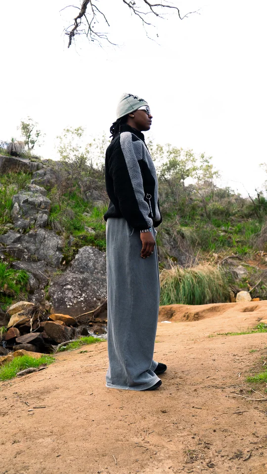 Model wearing a grey beanie, black jacket and wide grey trousers outdoors.