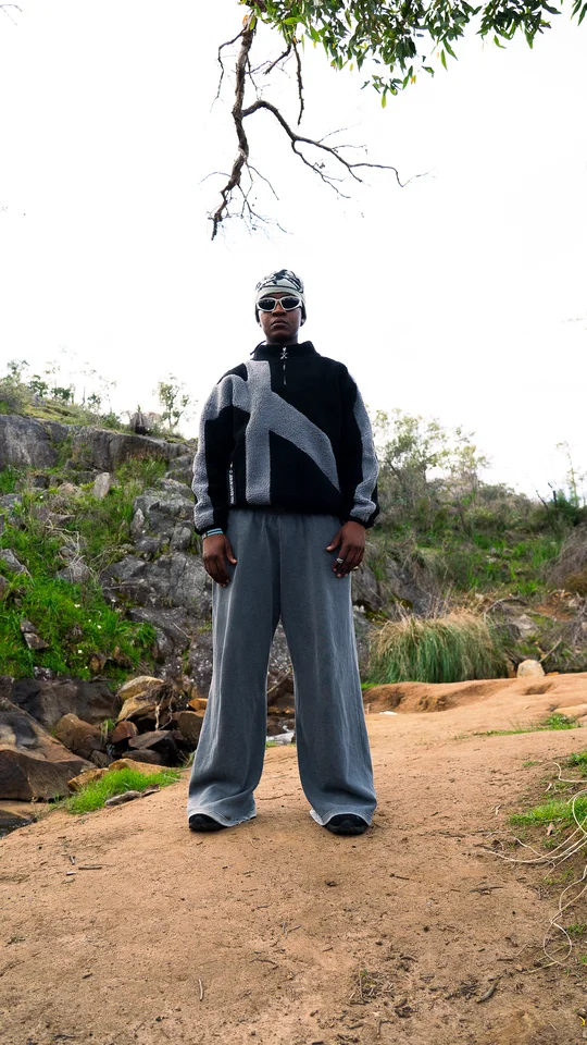 Model wearing a grey beanie, black jacket and wide grey trousers outdoors.