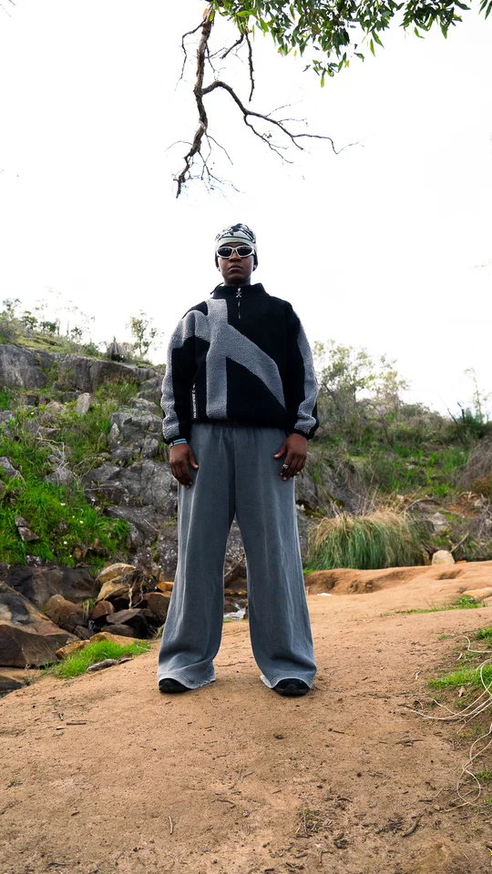 Model wearing a grey beanie, black jacket and wide grey trousers outdoors.