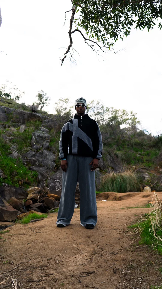 Model wearing a grey beanie, black jacket and wide grey trousers outdoors.