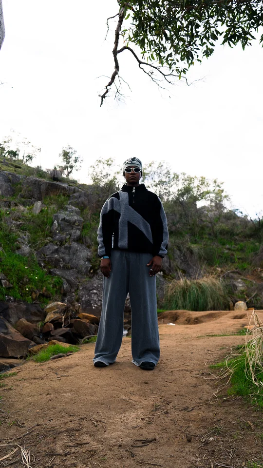 Model wearing a grey beanie, black jacket and wide grey trousers outdoors.