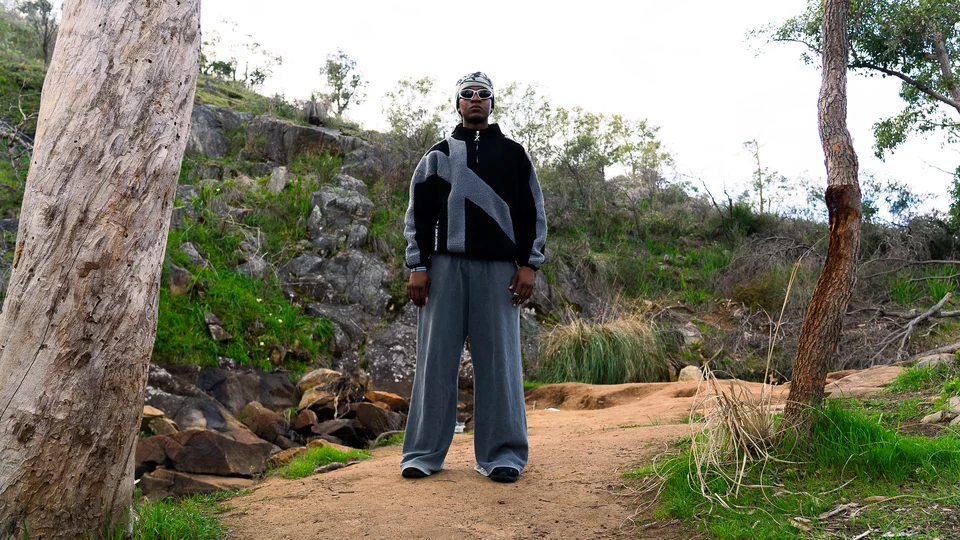 Model wearing a grey beanie, black jacket and wide grey trousers outdoors.