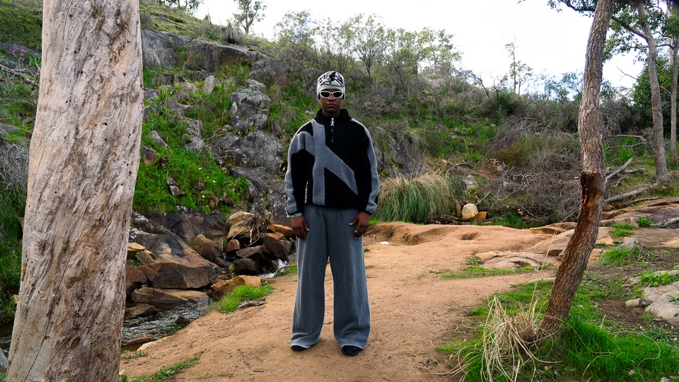Model wearing a grey beanie, black jacket and wide grey trousers outdoors.