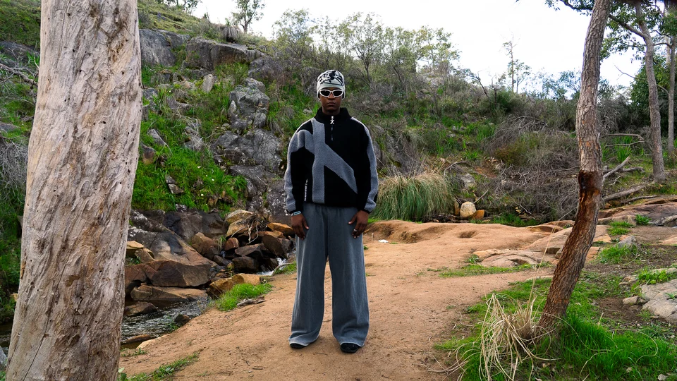 Model wearing a grey beanie, black jacket and wide grey trousers outdoors.